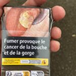 "Smoking provokes mouth and neck cancer," in French. The figure shows tongue cancer. A graphic cigarette pack thrown on the ground in Hoenheim, north of Strasbourg, France. Most cigarettes come from Germany, where they are cheaper because they are taxed less than in France. Germany is just over the bridge from Strasbourg. There are a lot of smokers in France, who seem undeterred by the images, and who also disproportionately throw litter on the ground. Was tut man?
